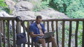 Casual businessman working with laptop at waterfall. The man, who is on vacation by the waterfall in nature, works by taking his laptop with him. video