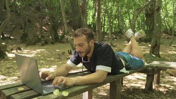 Young man working with laptop in forest. Young man lying on a bench works with a laptop. video