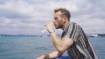 Bebiendo agua en un caliente día. el joven hombre, quien es abrumado por el calor por el mar en verano, bebidas su agua. video