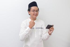 Portrait of young Asian muslim man raising his fist, celebrating winning game or getting good news on his mobile phone. Isolated image on white background photo