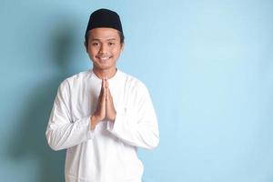 Portrait of Asian muslim man in white koko shirt with skullcap showing apologize and welcome hand gesture. Isolated image on blue background photo