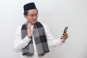 Portrait of smiling young Asian muslim man taking picture of himself or doing video call, saying hi and waving his hand. Isolated image on white background photo