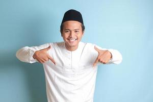 Portrait of young Asian muslim man showing product and pointing down with his hand and finger to the side. Isolated image on blue background photo