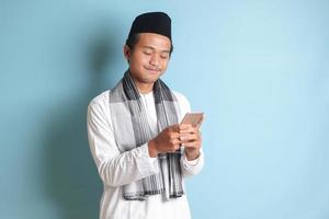 Portrait of young Asian muslim man holding and touching mobile phone with smiling expression on face. Isolated image on blue background photo