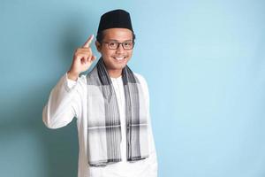 Portrait of young Asian muslim man pointing up his forefinger and reminding something to do. Advertising concept. Isolated image on blue background photo