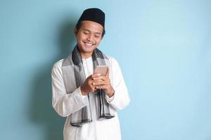 Portrait of young Asian muslim man holding and touching mobile phone with smiling expression on face. Isolated image on blue background photo