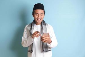 Portrait of young Asian muslim man holding and touching mobile phone with smiling expression on face. Isolated image on blue background photo