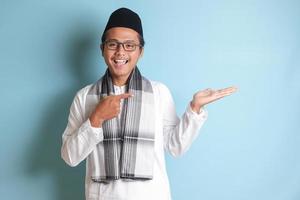 Portrait of young Asian muslim man showing product and pointing with his hand and finger to the side. Isolated image on blue background photo