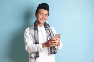 Portrait of young Asian muslim man holding and touching mobile phone with smiling expression on face. Isolated image on blue background photo