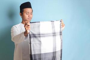 Portrait of Asian muslim man in white koko shirt folding and tidy up his sarung . Isolated image on blue background photo