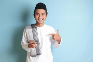 Portrait of young Asian muslim man holding mobile phone with smiling expression on face while pointing finger to the side. Isolated image on blue background photo