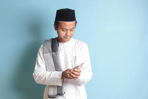 Portrait of young Asian muslim man holding and touching mobile phone with smiling expression on face. Isolated image on blue background photo