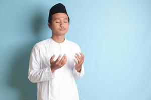 retrato de asiático musulmán hombre en blanco koko camisa con casquete Orando formalmente con su manos aumentó. aislado imagen en azul antecedentes foto