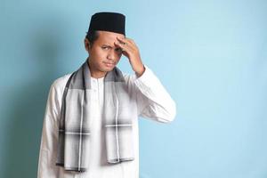 Portrait of attractive Asian muslim man in white shirt having a migraine, touching his temple. Headache disease concept. Isolated image on blue background photo