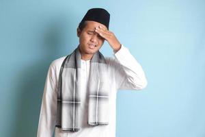 retrato de atractivo asiático musulmán hombre en blanco camisa teniendo un migraña, conmovedor su templo. dolor de cabeza enfermedad concepto. aislado imagen en azul antecedentes foto