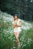 hermosa joven mujer cosecha blanco flores silvestres en el antecedentes de un bosque paisaje en verano. retrato de un amable contento mujer en un salvaje campo, disfrutando naturaleza. natural belleza modelo foto