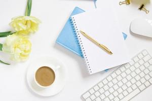 Notepad with a gold pen, yellow tulip flowers, a cup of coffee, a keyboard and a cup of coffee on a white background. Stylish workplace of a blogger or freelancer photo