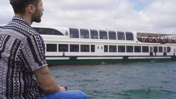 de Mens op zoek Bij de zee. Mens zittend Aan de strand looks uit naar zee en wit passagier veerboot in de achtergrond. video