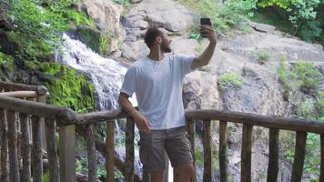 Talking on the phone by the waterfall in nature. Handsome young man video chatting on phone in forest.