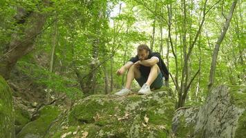 nachdenklich jung Mann im das Wald ist Sitzung auf das Felsen. das jung Mann ist nachdenklich und traurig. video