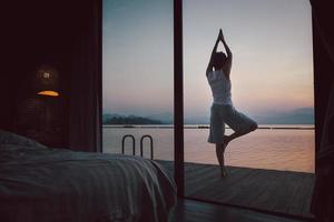 Woman doing yoga exercise on wooden balcony in bed room at sunrise. photo