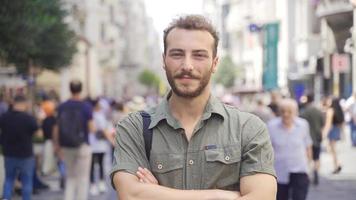 giovane uomo in piedi nel il mezzo di il folla guardare a il telecamera. contento sorridente giovane uomo guardare a telecamera nel affollato strada. video