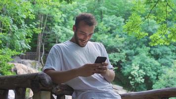 Texting on the phone at the waterfall in the forest. Young man standing in forest in front of waterfall and texting on his phone. video