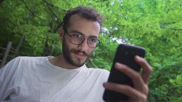 giovane uomo guardare a Telefono sotto alberi. uomo nel foresta parco guardare a Telefono. video