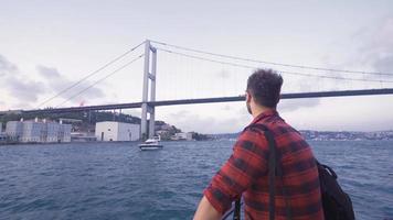 The man watching the bridge from the ferry. The man advancing on the ferry watches the vehicles passing over the bridge in the Bosphorus. video