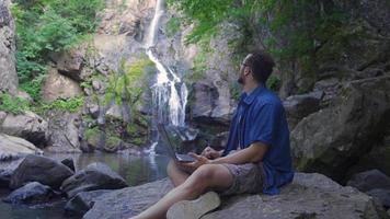 Working With Laptop In A Rocky Valley. Man resting in nature using laptop. Close-up, Beautiful flowing waterfall from behind. video