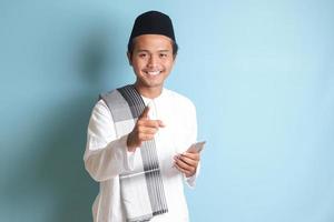 Portrait of young Asian muslim man holding mobile phone with smiling expression on face while pointing finger to the side. Isolated image on blue background photo