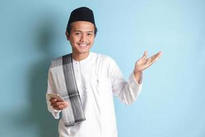 Portrait of young Asian muslim man holding mobile phone with smiling expression on face while pointing finger to the side. Isolated image on blue background photo