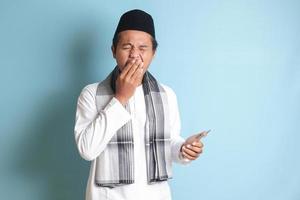 Portrait of young Asian muslim man holding mobile phone with while yawning with hand covering his mouth. Isolated image on blue background photo