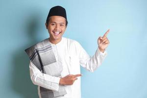 Portrait of young Asian muslim man showing product and pointing with his hand and finger to the side. Isolated image on blue background photo