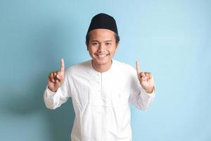 Portrait of young Asian muslim man showing product and pointing up with his hand and finger to the side. Isolated image on blue background photo