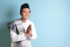 Portrait of Asian muslim man in white koko shirt with skullcap showing apologize and welcome hand gesture. Isolated image on blue background photo