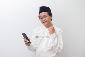Portrait of young Asian muslim man raising his fist, celebrating winning game or getting good news on his mobile phone. Isolated image on white background photo