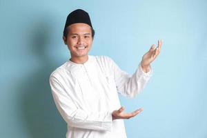 Portrait of young Asian muslim man showing product and pointing with his hand and finger to the side. Isolated image on blue background photo