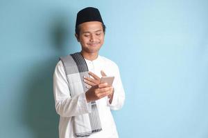 Portrait of young Asian muslim man holding mobile phone with smiling expression on face, touching heart, received good news. Isolated image on blue background photo