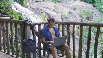 Holiday work. Young man at waterfall in nature works with laptop. video