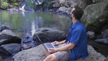 hombre trabajando con ordenador portátil a cascada. joven hombre sentado en un rock en naturaleza trabajando con computadora portátil, acecho cascada detrás a él. video