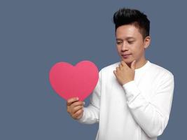 Handsome man holding red heart shape with finger on chin photo