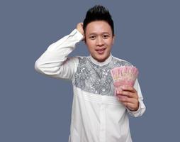 Young man holding Indonesian one hundred thousand rupiah with smiling face expression and hand scratching head photo