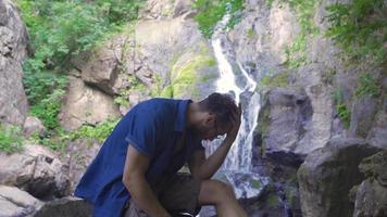 verdrietig Mens in natuur. eenzaam en verdrietig Mens zittend Bij waterval in Woud en denken. video