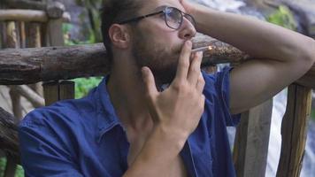 frustriert jung süchtig Rauchen Zigarette draußen. jung Mann Sitzung durch das Wasserfall Rauchen ein Zigarette. video