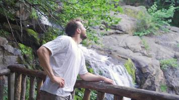 Tourist man is resting at the waterfall. Young tourist spending time in relaxing cool waterfall looking at waterfall. video