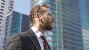 Young Businessman talking on the phone between big buildings. Businessman talking on the phone on a sunny day looks at his watch and continues talking on the phone. video