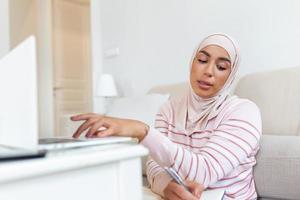 Muslim young women wear hijab using typing and checking order the customer address on the computer laptop at home morning. Concept business small and shop online photo