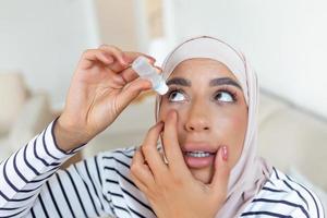 Arábica mujer con hijab utilizando ojo gota, goteante ojo lubricante a tratar seco ojo o alergia, enfermo mujer tratando globo ocular irritación o inflamación mujer sufrimiento desde irritado ojo, óptico síntomas foto