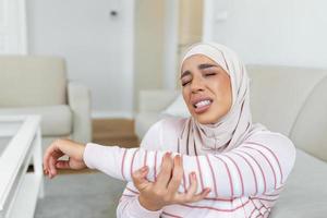 joven Arábica musulmán mujer con hijab es sentado a hogar en su sofá y conmovedor el su codo mientras sufrimiento desde codo dolor foto
